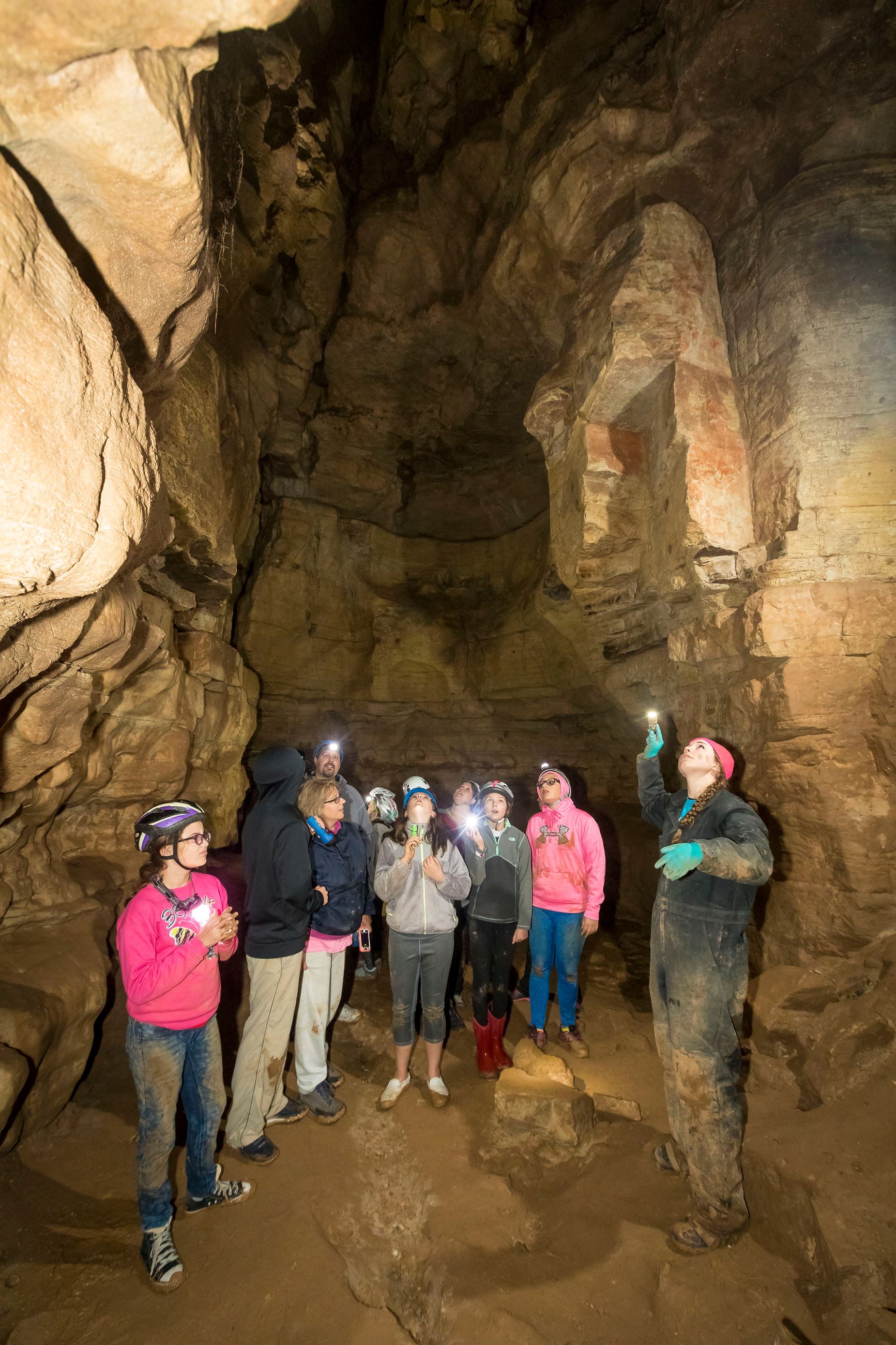 An instructor and a group of teenage girls standing in a large cave. They are all looking up.